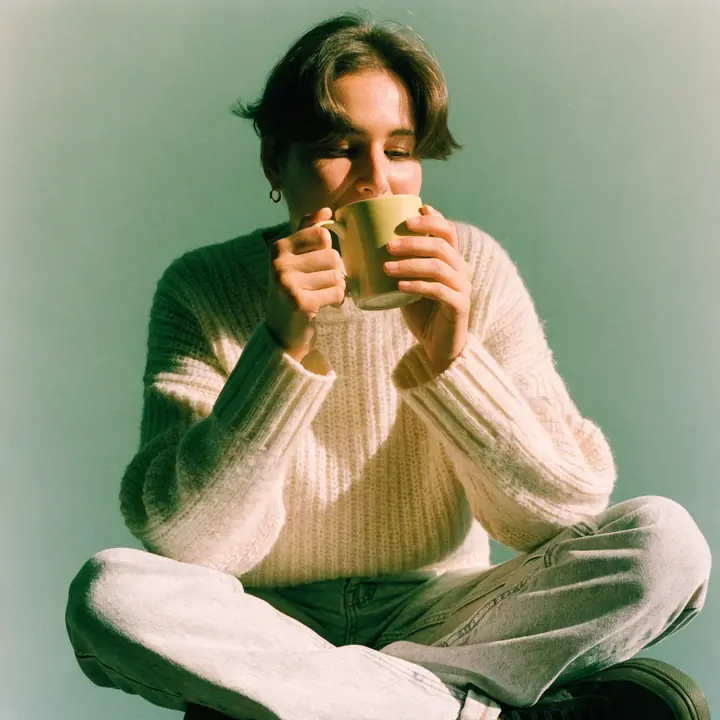 A person sitting cross-legged in a knitted jumper drinking from a big mug against a green wall