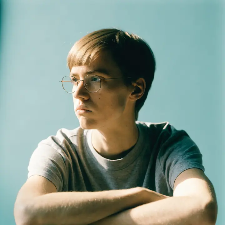 A person in round glasses and a plain grey t-shirt with arms folded, looking away, on a pale blue backdrop